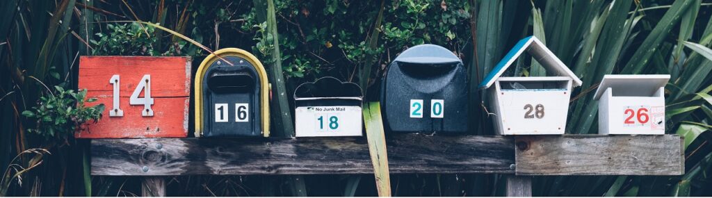 six assorted-color mail boxes on a wooden bench in front of shrubbery
