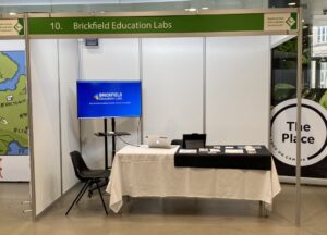 Brickfield Education Labs exhibition stand at ALTC22 - with apresentation playing on a Big Screen, a table with brochures onit.